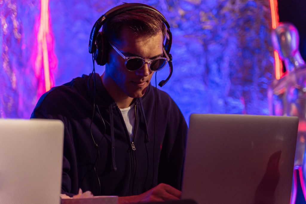 A man wearing sunglasses and headset works on a laptop in a colorful setting.